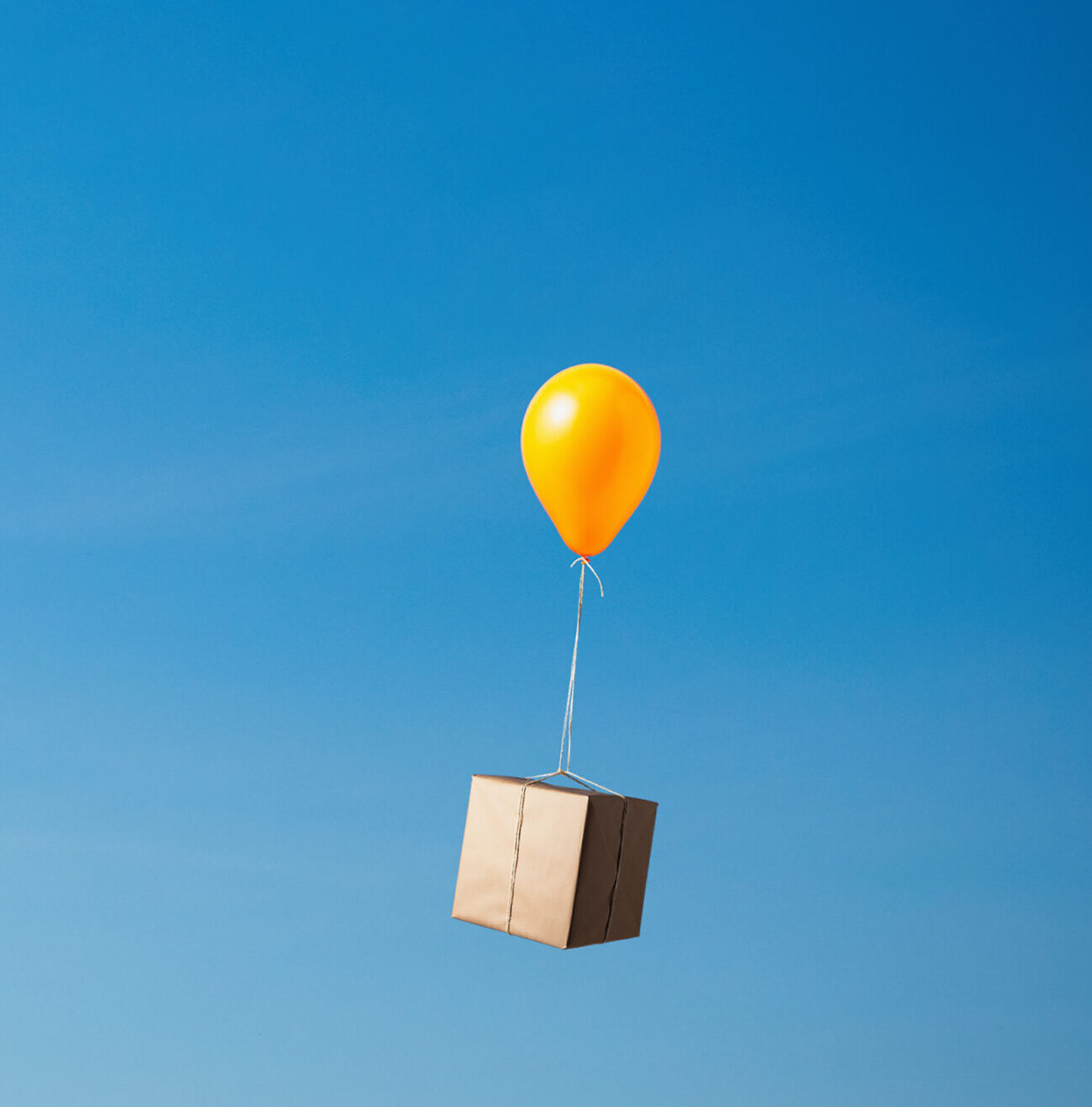 One yellow helium balloon carrying parcel wrapped in brown paper tied with string over grassy field, against blue sky